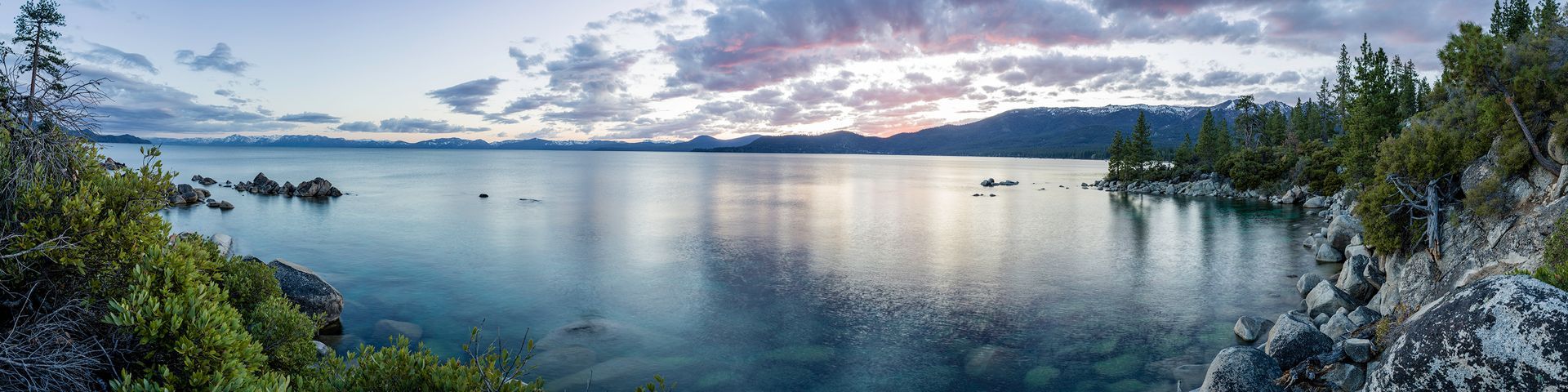 Calm lake at sunset with rocky shoreline, trees, and dramatic clouds reflecting on the water
