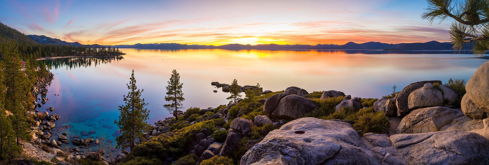 Snowy mountain lake at sunset with pine trees, rocky shoreline, and colorful sky reflections