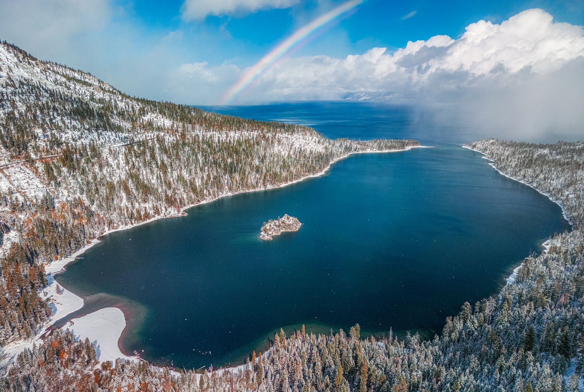 Snow-covered mountains framing a deep blue lake with a small island under cloudy skies