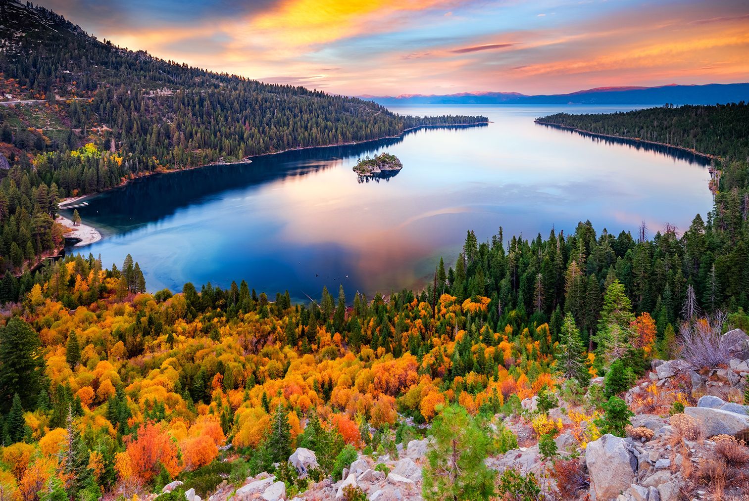 Colorful autumn forest framing a calm blue lake at sunset with mountains in the distance