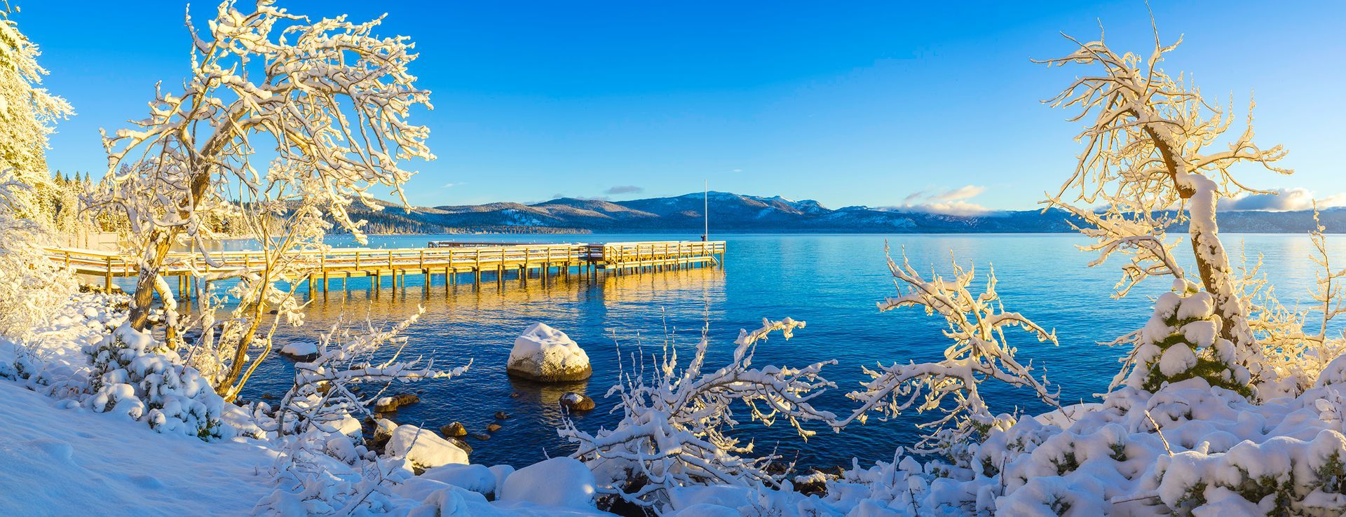 Snowy trees framing a bright blue lake with a small pier under a clear winter sky