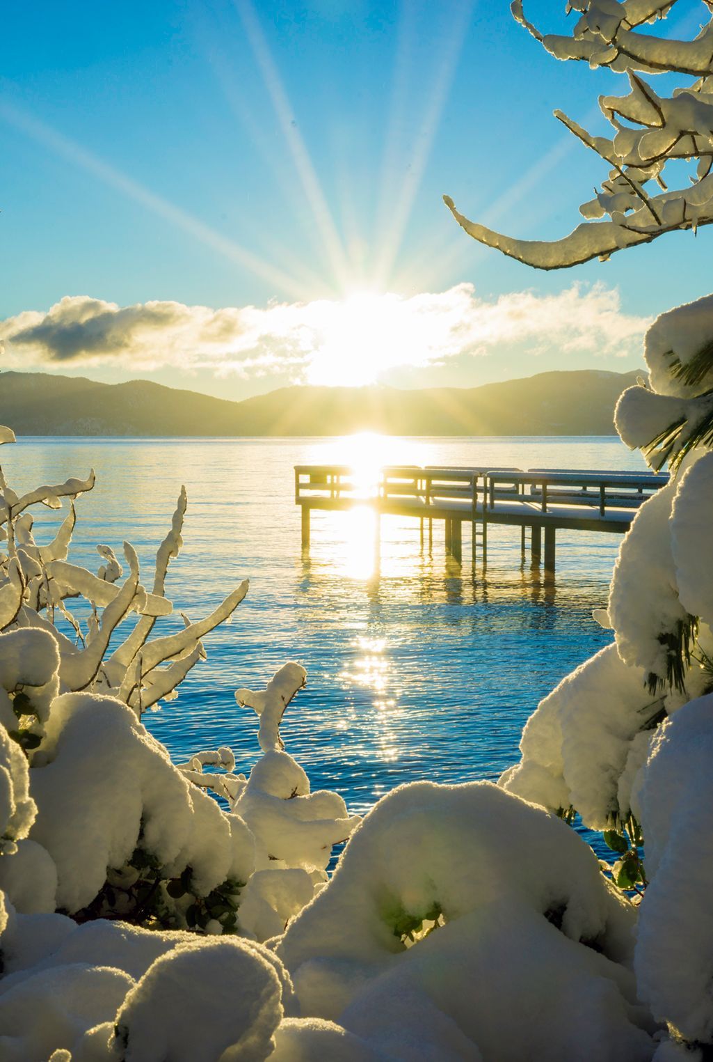 Snow-covered lakeside dock at sunrise with bright sun reflecting on calm blue water