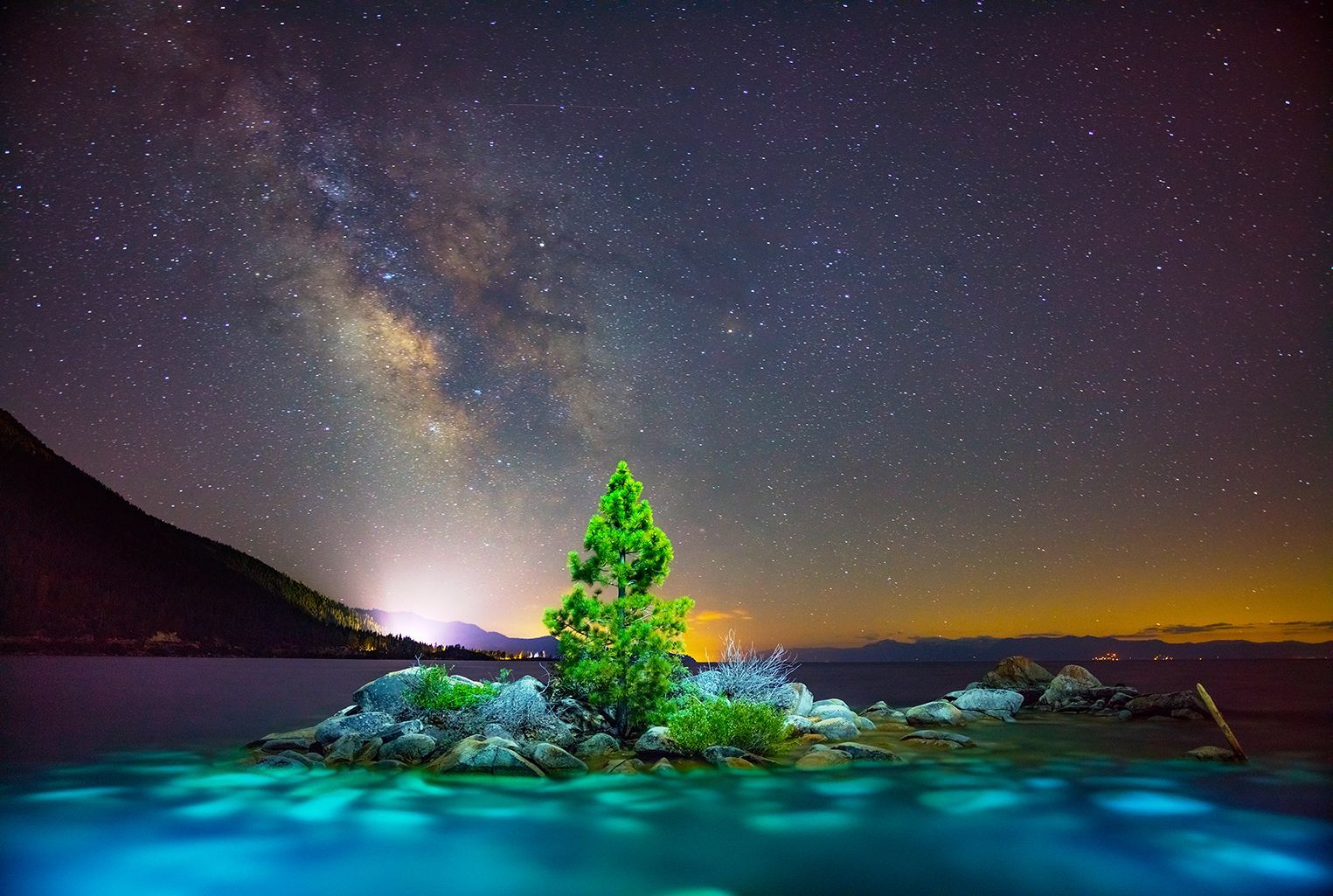 Milky Way over a glowing turquoise lake with a lit tree on the shore at night