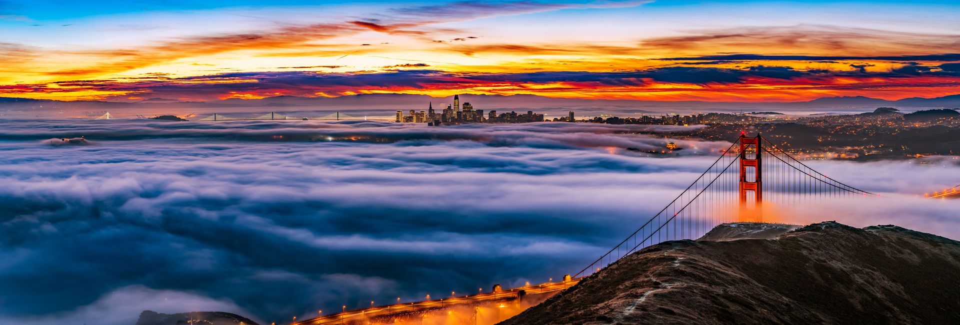 Golden Gate Bridge at sunrise above fog, with a vivid orange and purple sky.