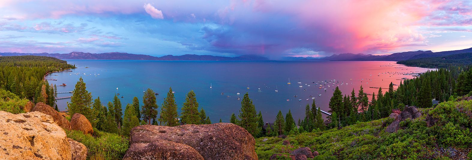Panoramic lake at sunset with pink clouds, blue sky, rocky shoreline, and evergreen forest