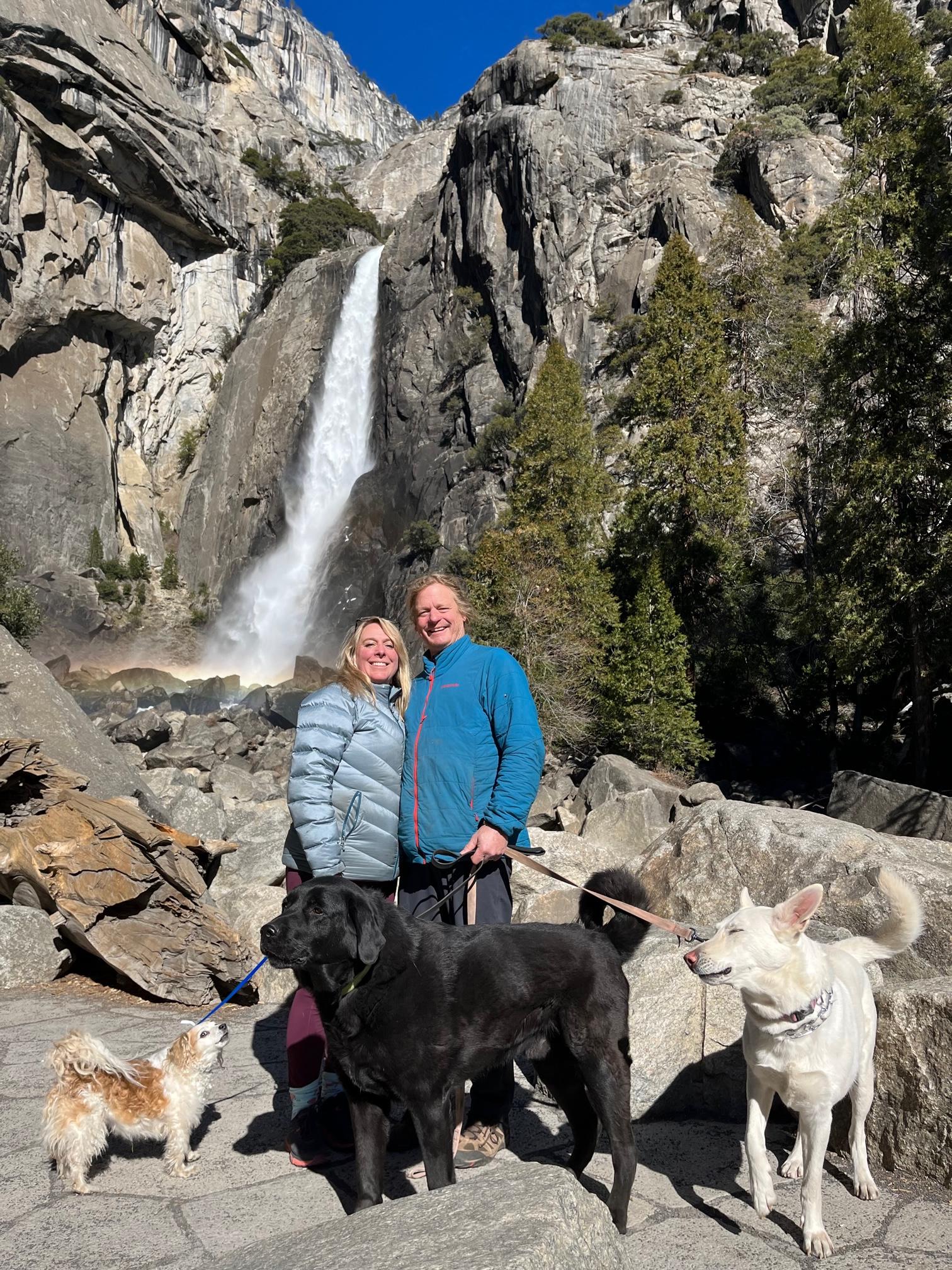 Two people and three dogs posing before a tall waterfall in a rocky mountain setting