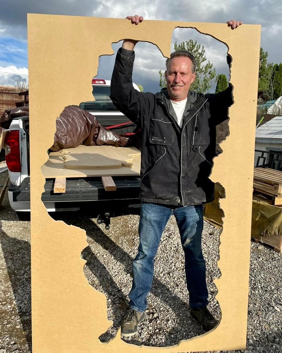 Man posing in a yellow cutout board outdoors, holding a top edge with one arm raised.