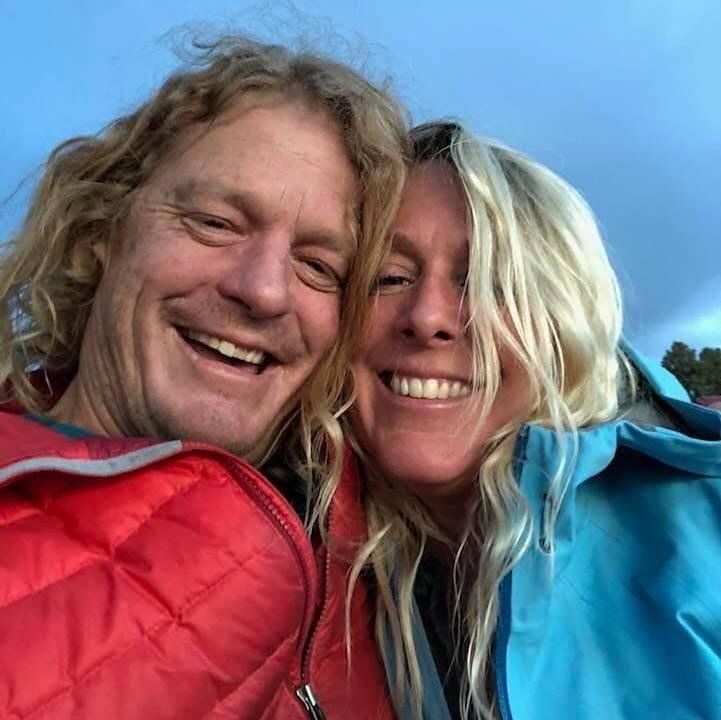 Two smiling people in red and blue jackets posing close together outdoors under a blue sky