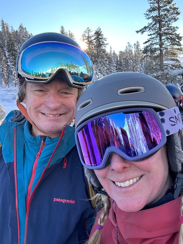 Two people in ski goggles smiling outdoors in snowy mountains at sunset