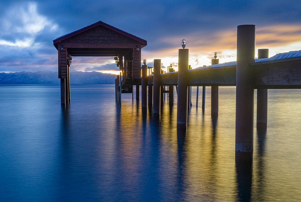 Wooden pier and gazebo at sunset over calm blue water, with silhouetted posts and cloudy sky.