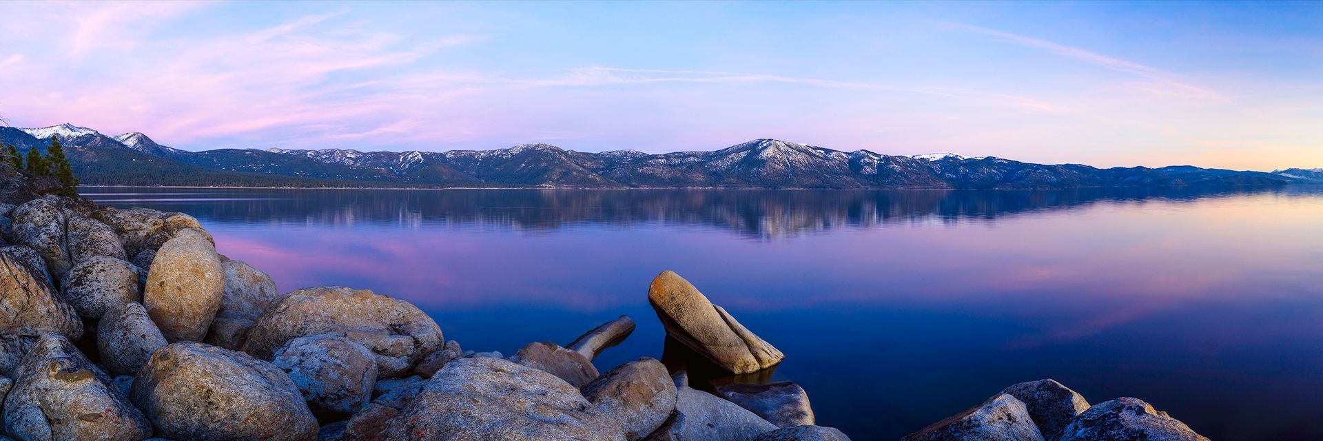 Rocky lakeshore at dawn with calm blue water, distant trees, and pink-purple sky