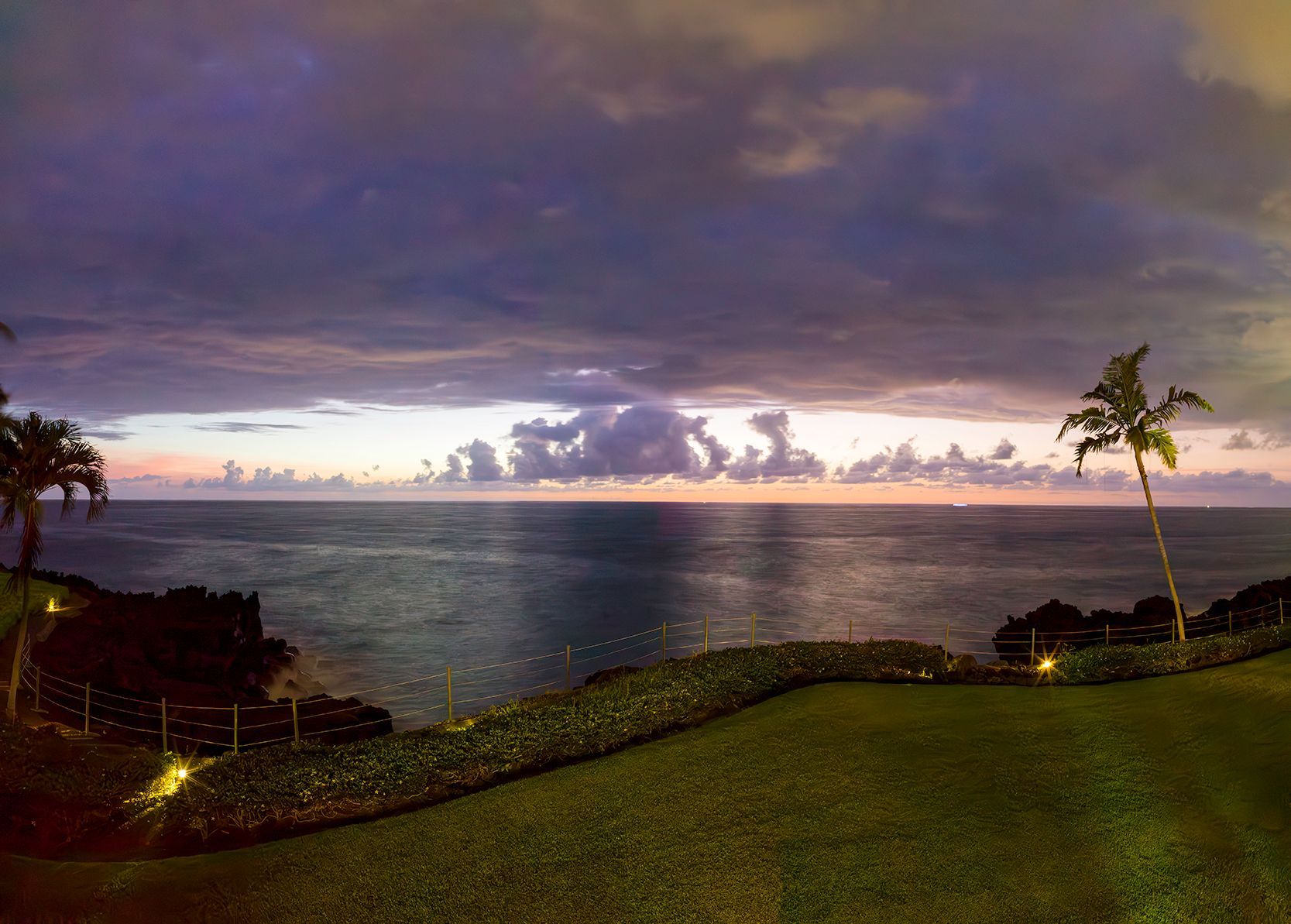 Sunset over the ocean with dark clouds, a lit grassy shoreline, and a lone palm tree.