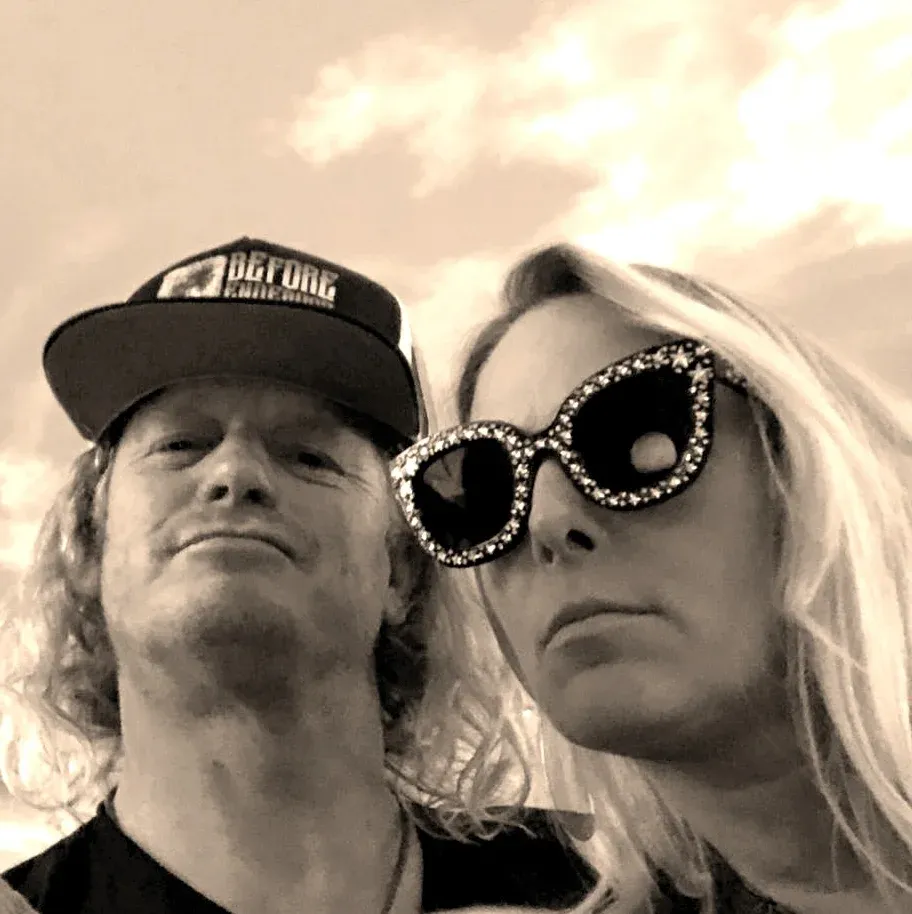 Two people wearing hats and sunglasses posing outdoors under a cloudy sky