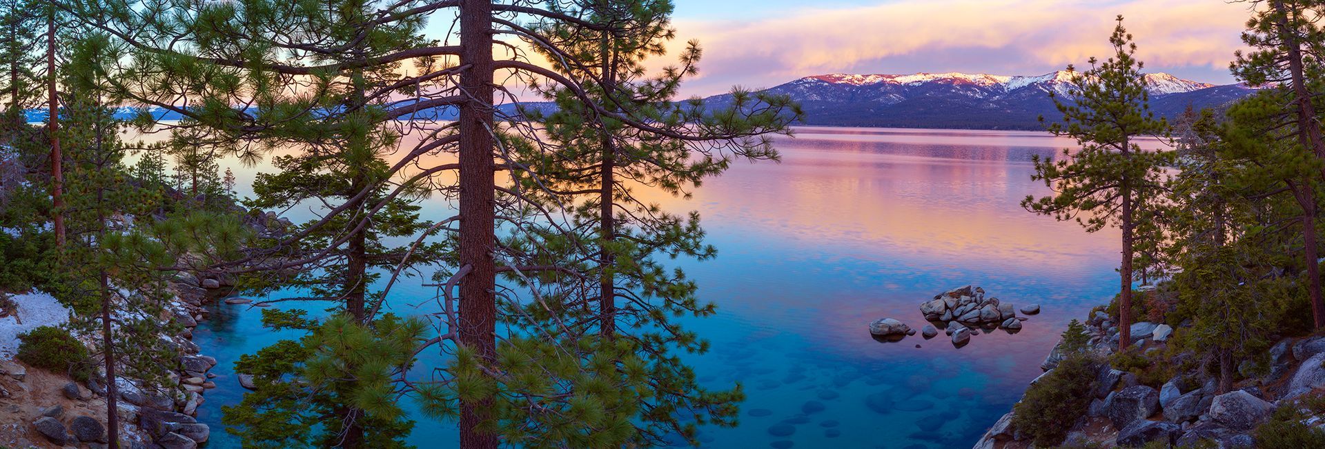 Sunset over a blue lake framed by pine trees and distant snow-capped mountains