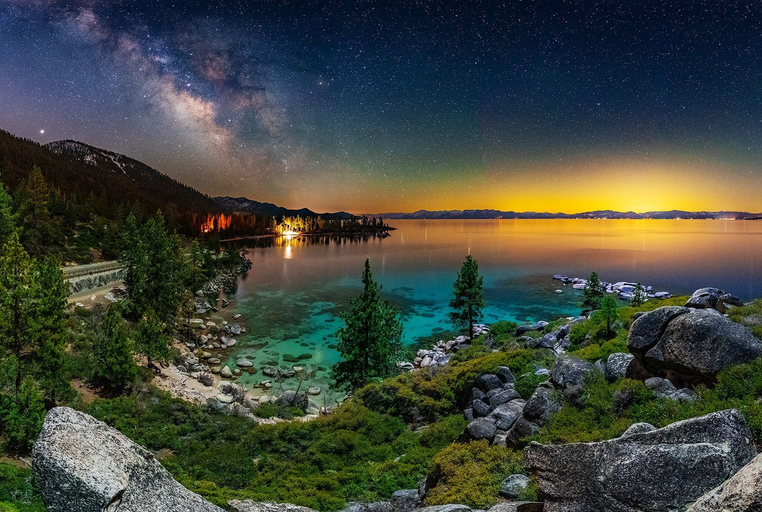 Night sky over a turquoise cove with rocky shore, trees, and the Milky Way above a glowing horizon