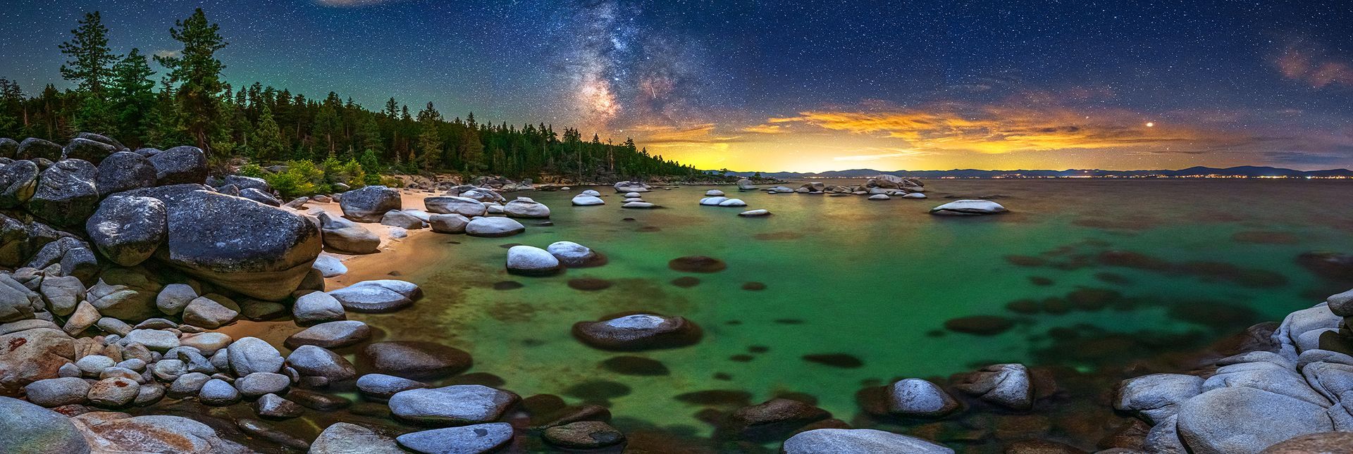 Rocky lakeshore at dusk with green water, pine trees, and a glowing orange horizon under a starry sky