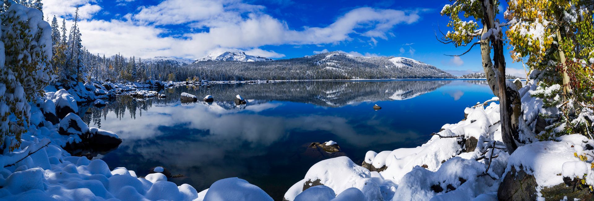 Snowy mountain lake under a blue sky, with snow-covered trees and rocks in the foreground.