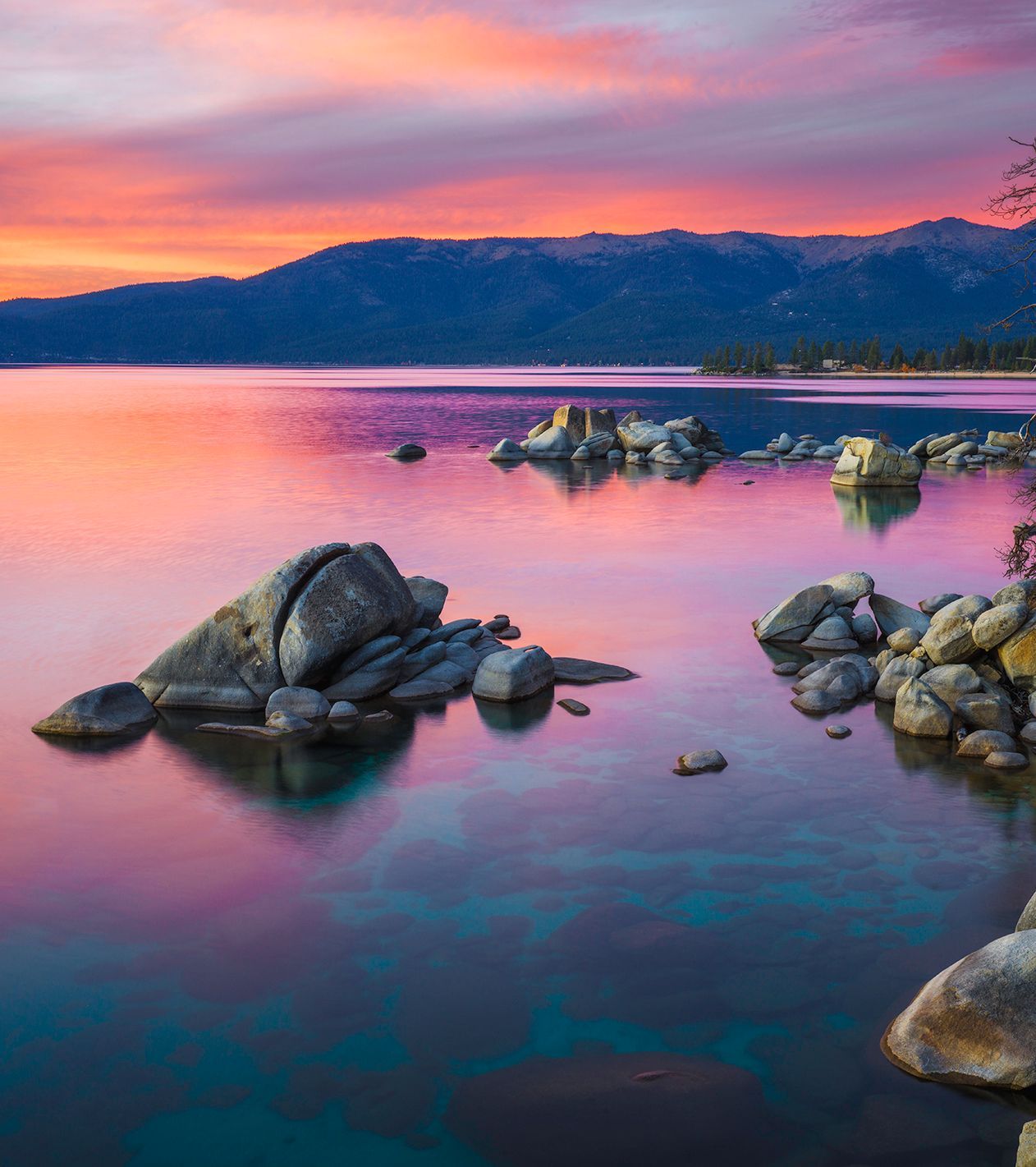 Rocky lakeshore at sunset with pink reflections and distant mountains beneath a colorful sky