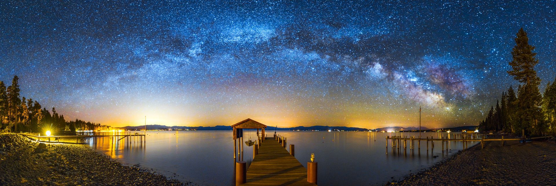 Milky Way over a calm lake at dusk, with a wooden dock and glowing lights along the shore