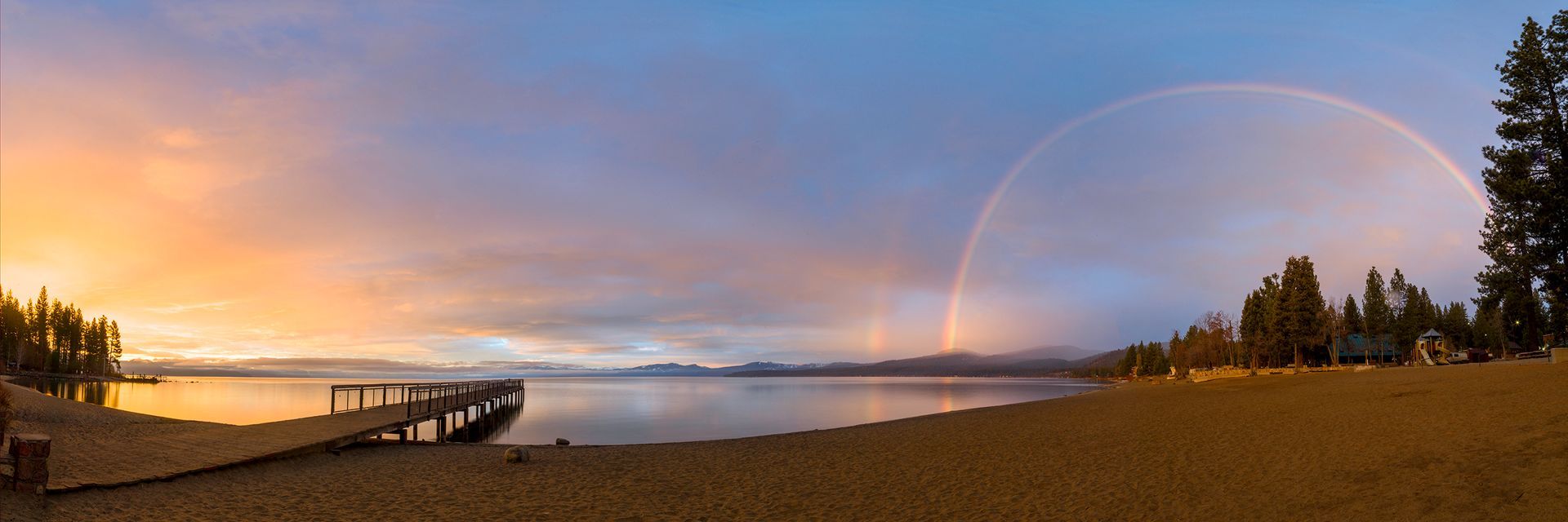 Sunset over a beach with a pier, calm water, and a faint rainbow in the sky