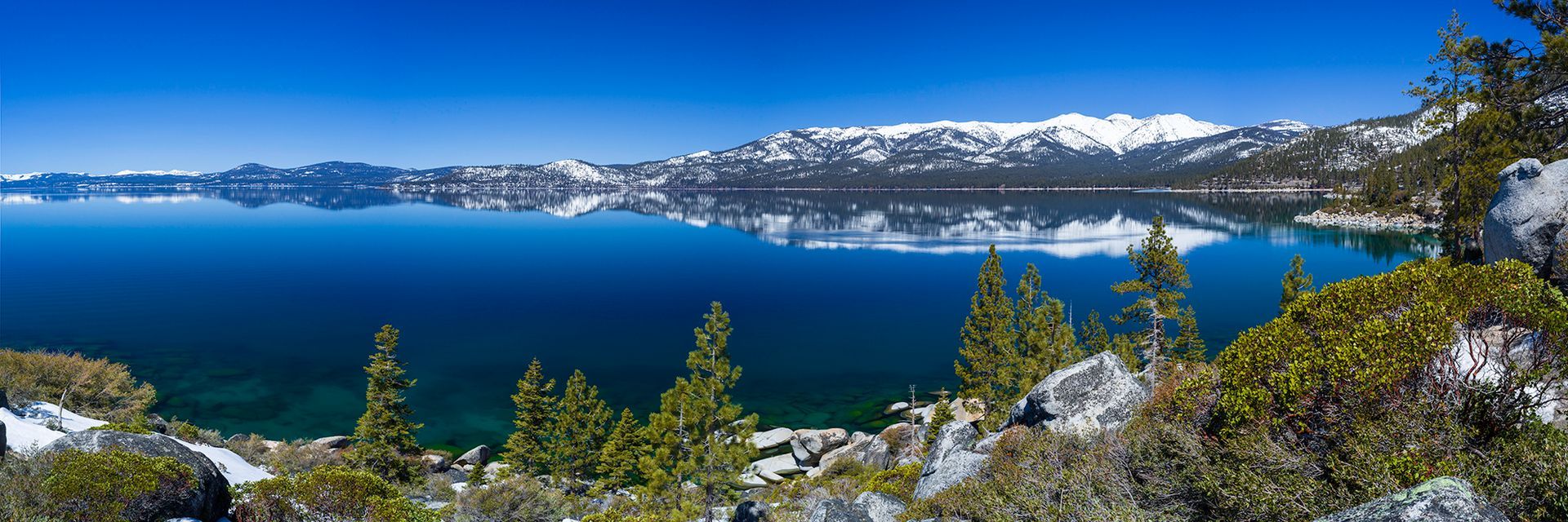 Snowy mountain lake with deep blue water, rocky shoreline, and evergreen trees under a clear sky