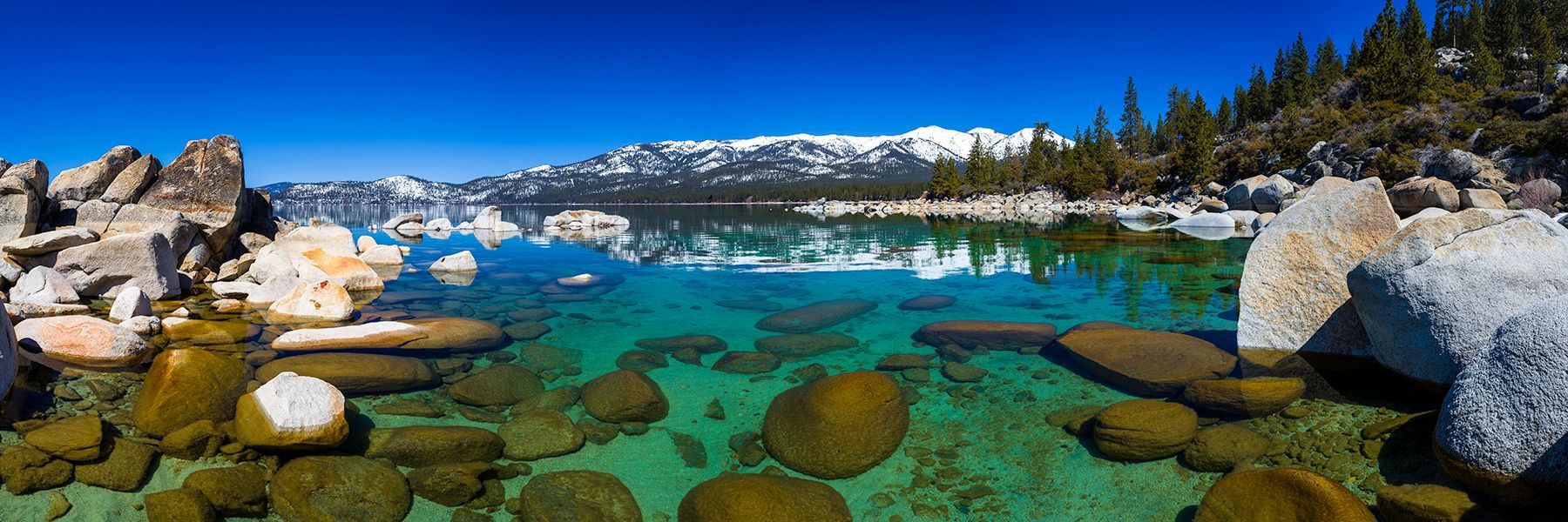 Turquoise alpine lake with rocks, pine trees, and snow-capped mountains under a clear sky