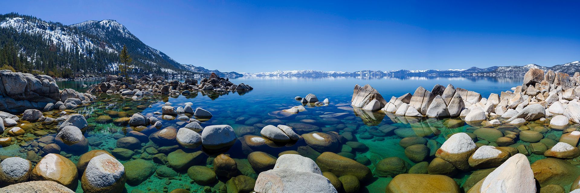 Rocky seaside cove with turquoise water, snow-dusted mountains, and a clear blue sky.