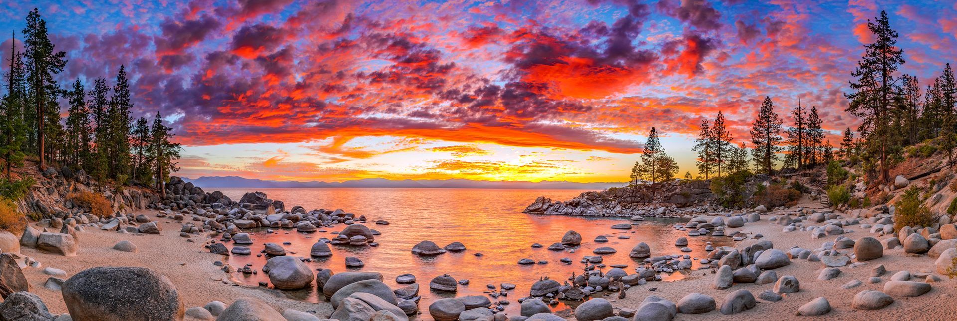 Rocky beach at sunset with vivid pink-orange sky reflected on calm water, framed by pine trees and boulders
