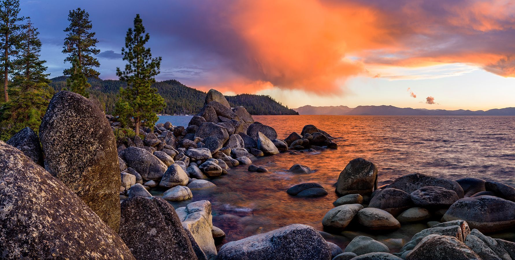 Rocky lakeshore at sunset with glowing orange clouds and pine trees along the water