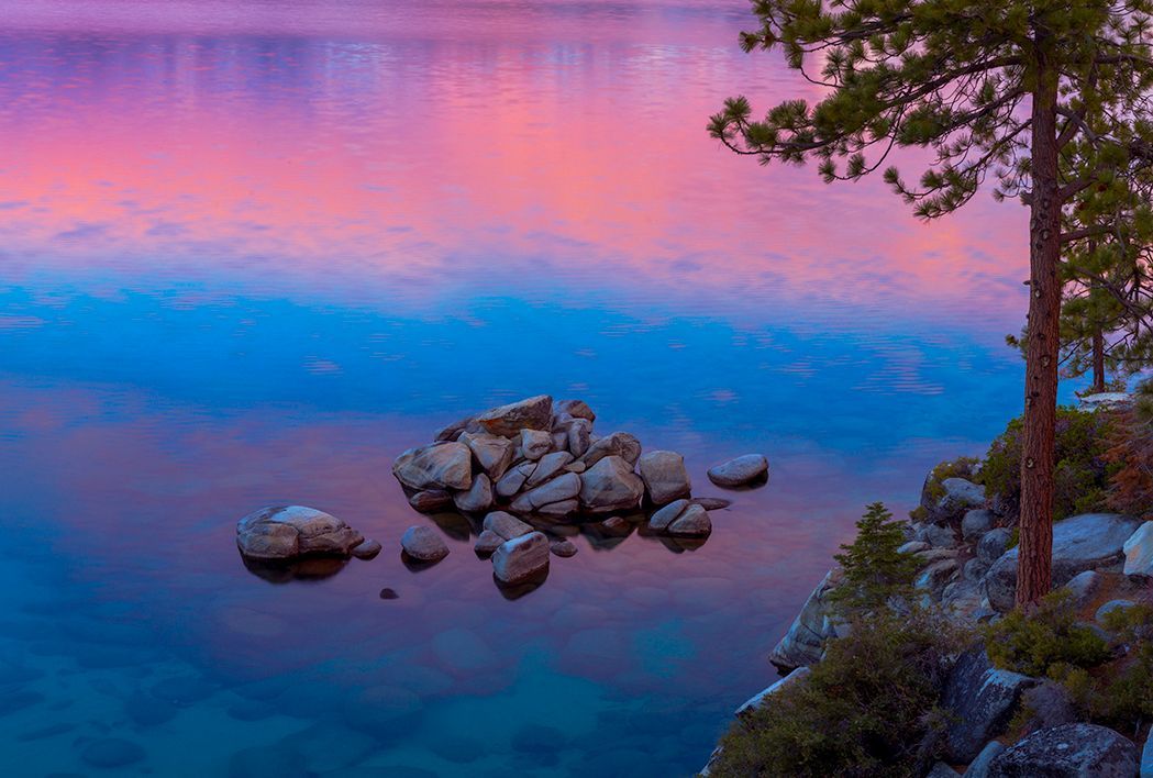 Pink-and-blue sunset over a calm lake with rocks and a tree-lined shore