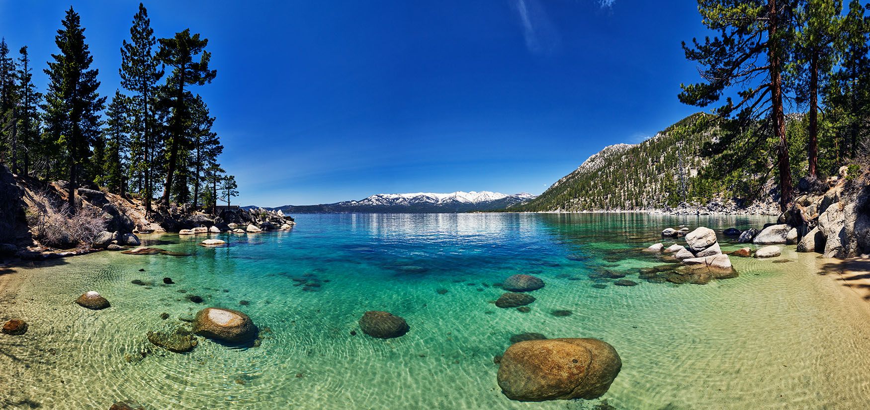 Clear turquoise alpine lake with rocky shore, pine trees, and snow-capped mountains under a bright blue sky