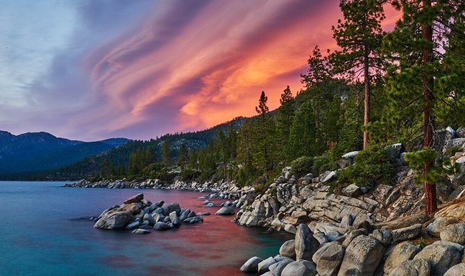 Rocky lakeshore at sunset with pine trees, mountains, and a pink-purple sky