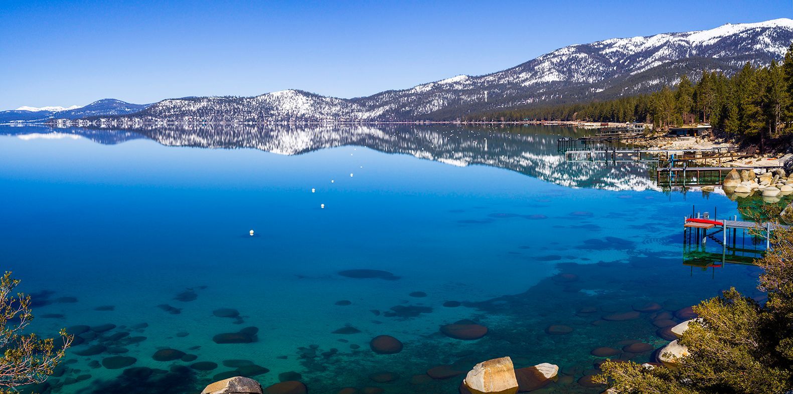 Snowy mountains overlook a calm blue lake with a rocky shoreline and a small dock