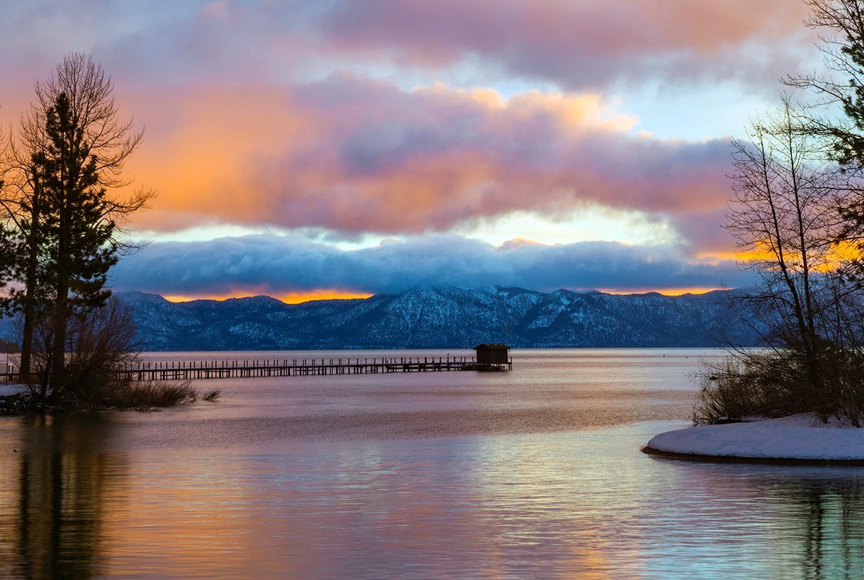 Snowy lakeshore at sunset with pink clouds, distant mountains, and a wooden pier over calm water