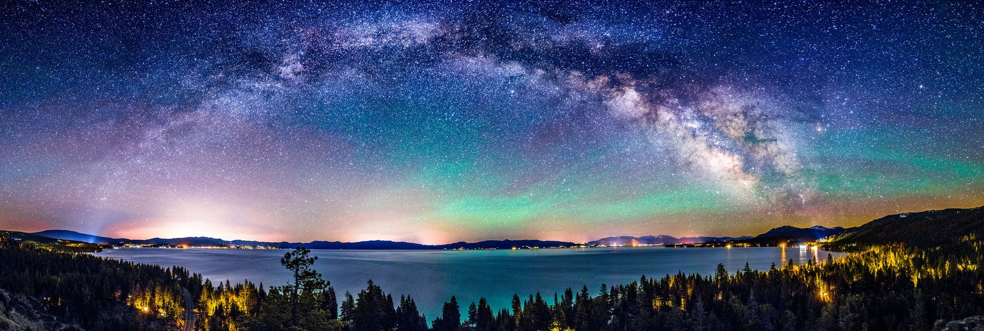 Panoramic night sky with Milky Way over a calm lake, distant mountains, and silhouetted trees in twilight colors
