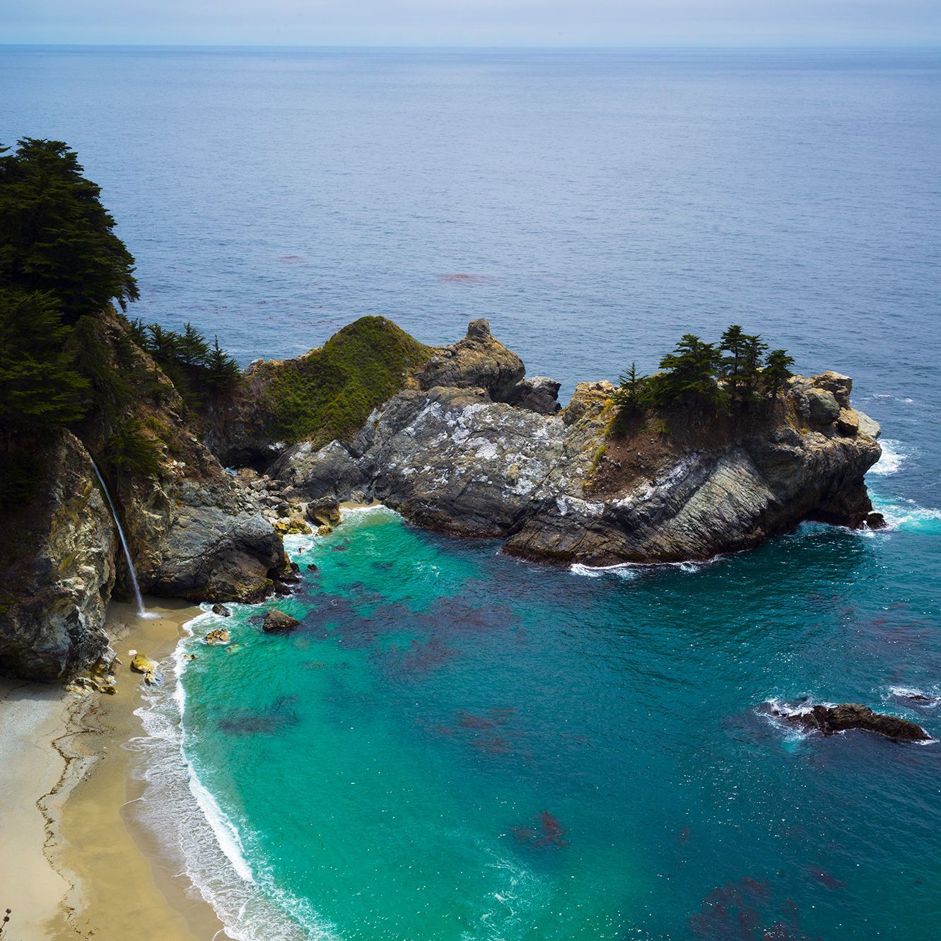 Aerial view of a turquoise cove with rocky cliffs and a small sandy beach under calm blue water