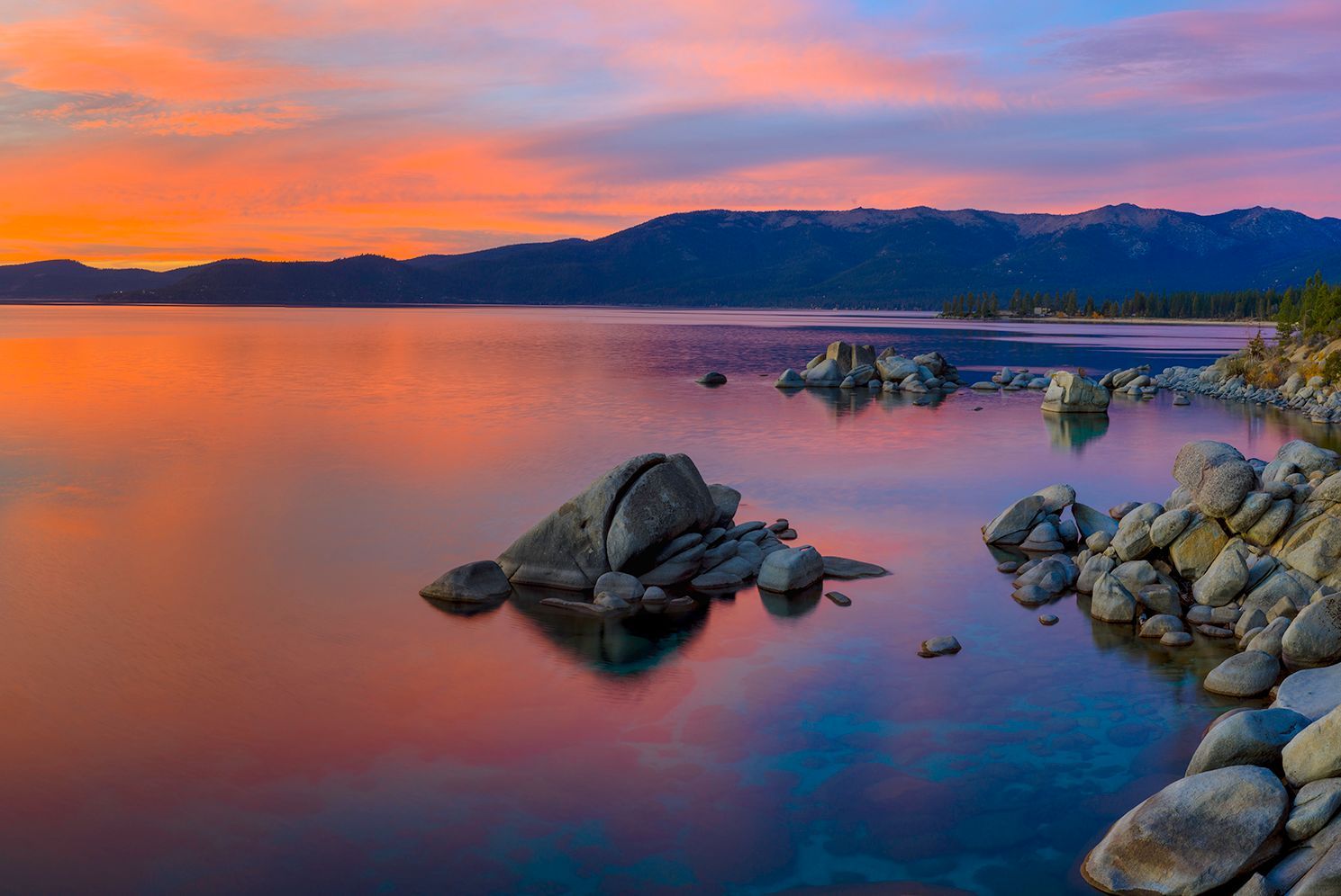 Sunset over a calm lake with rocks in the foreground and mountains silhouetted in the distance