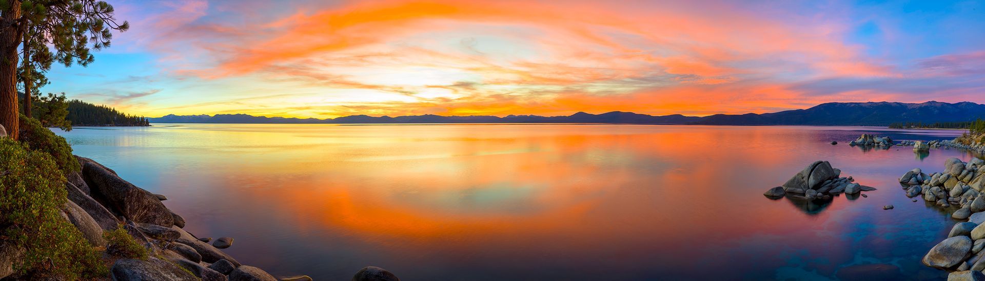 Sunset over a calm lake with orange and pink sky reflecting on still water, rocky shore in foreground