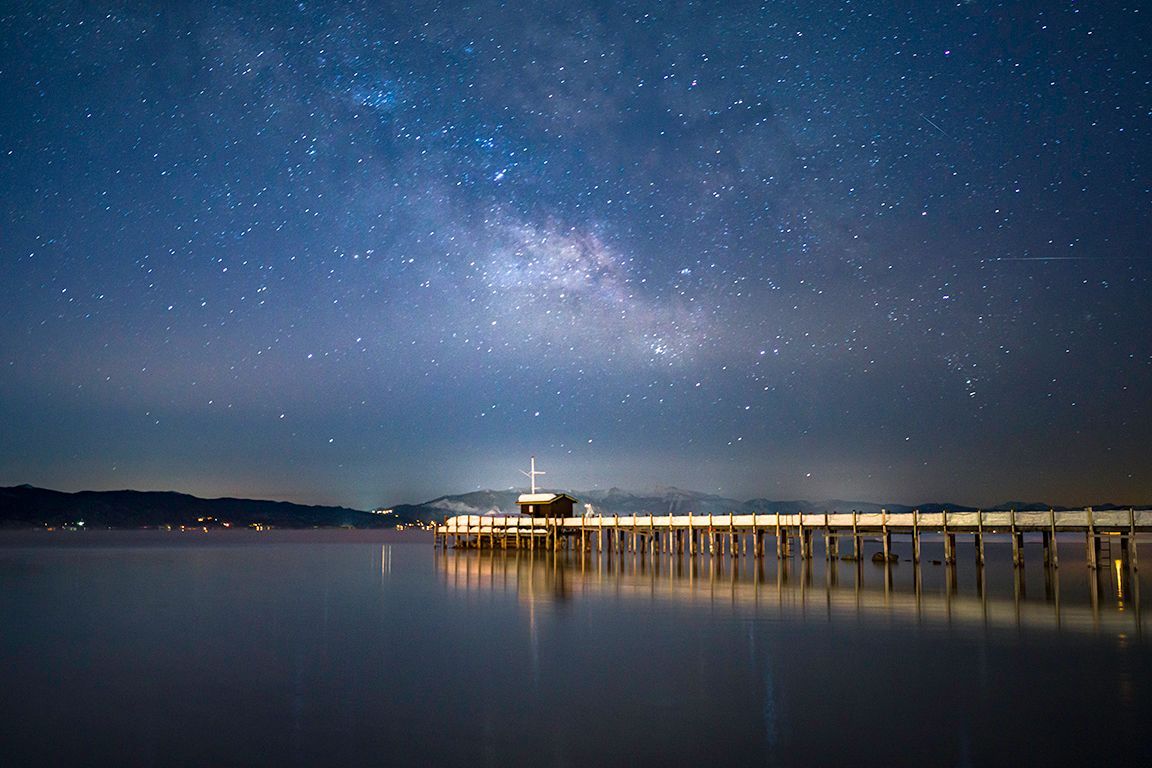 Night sky over a calm pier and lighthouse, with stars reflecting on still water