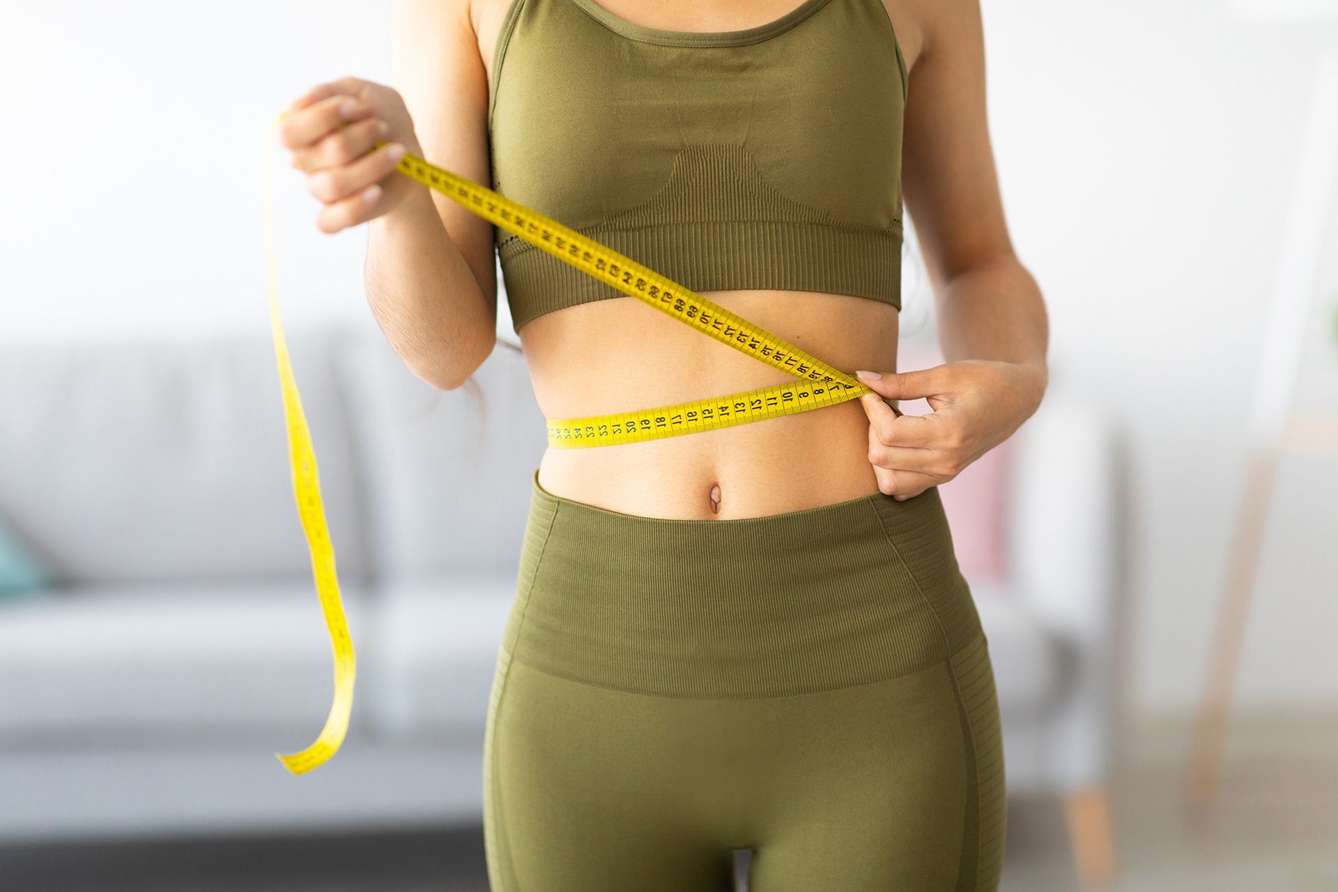 Cropped view of slim Indian woman measuring her waist with tape measure at home, closeup. Unrecognizable Asian lady checking result of weight loss diet or liposuction indoors