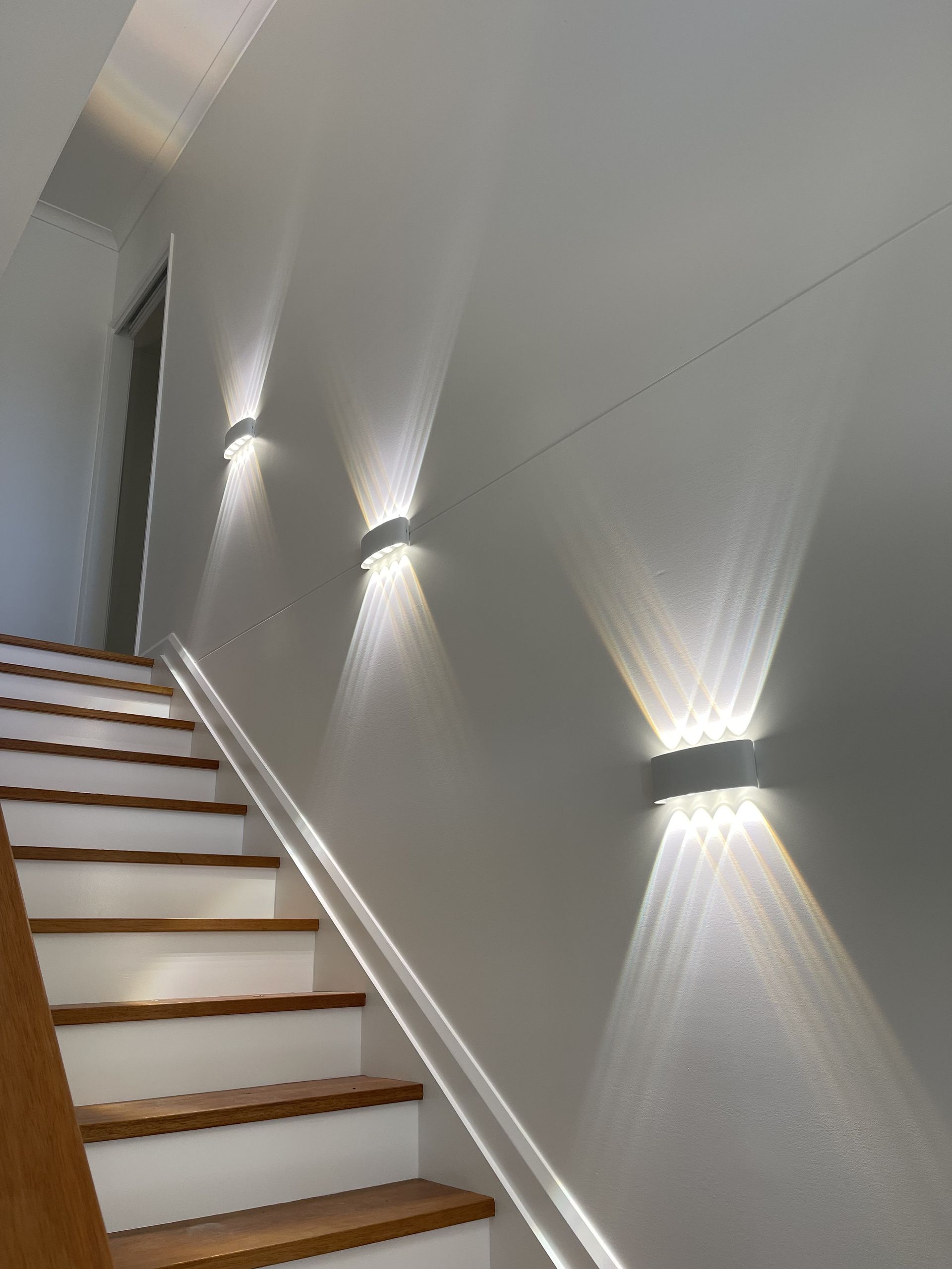 Modern stairwell with white walls, wooden steps, and wall-mounted lights casting patterns on the wall