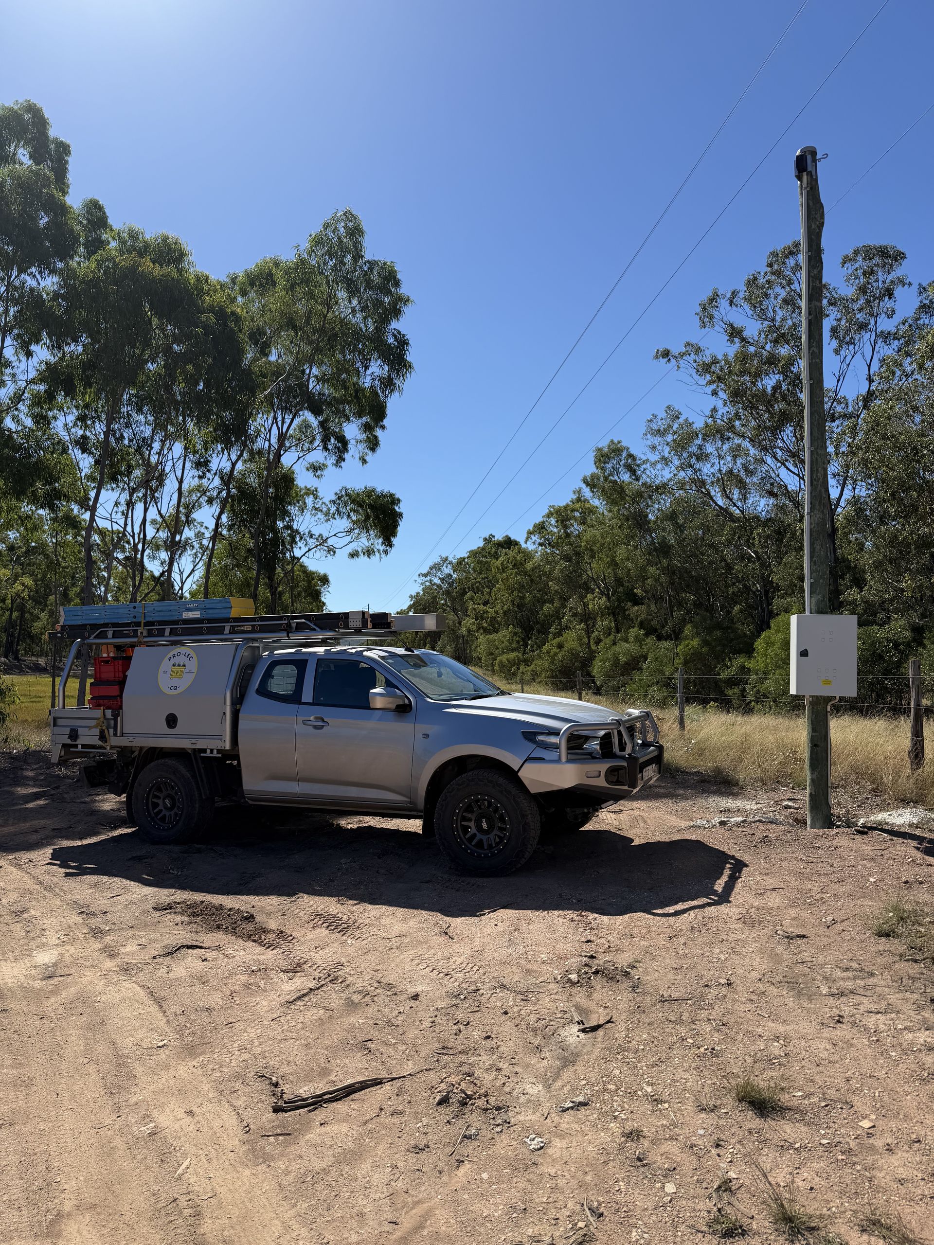 Silver utility truck on a dirt road beside trees and a utility pole under a clear blue sky
