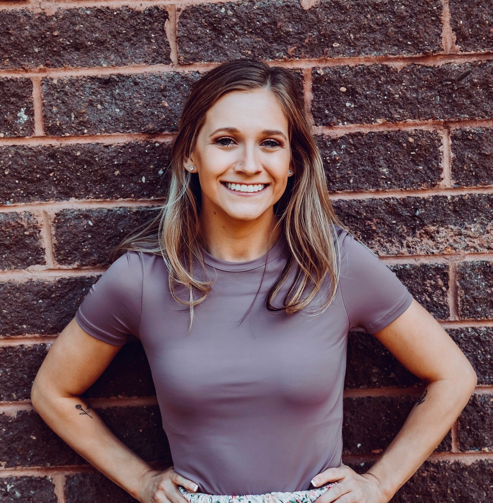 Woman with hands on hips smiles in front of a brick wall, wearing a purple shirt.