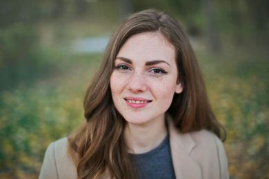 Woman with long brown hair and freckles, smiling outdoors in front of blurry foliage.