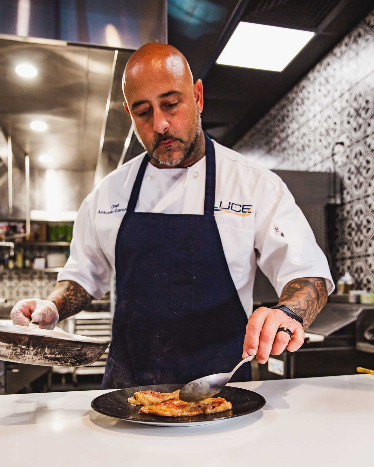 tony prepares a plate of food in a kitchen wearing an apron that says luce