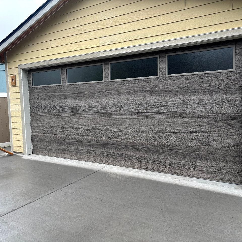 Gray garage door with small windows on a concrete driveway, under a beige house roofline