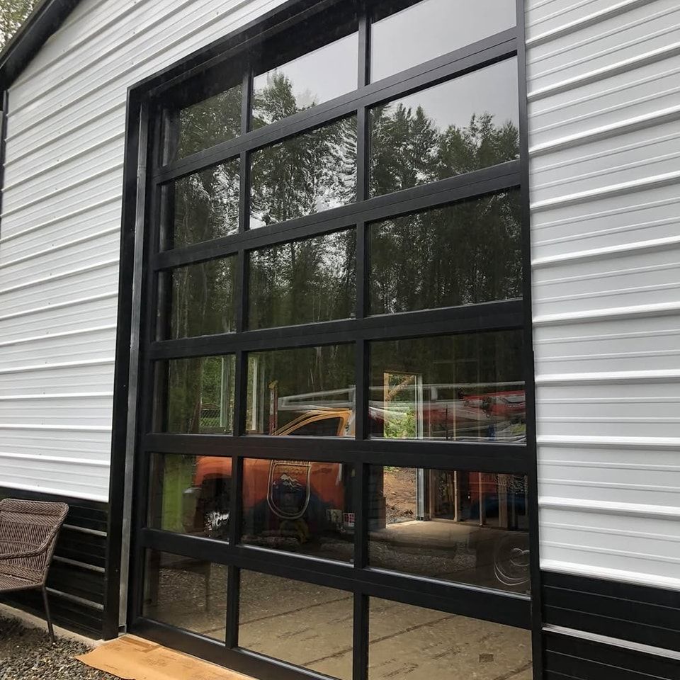 Modern black-framed glass garage door on a white house exterior with reflections of trees and a vehicle