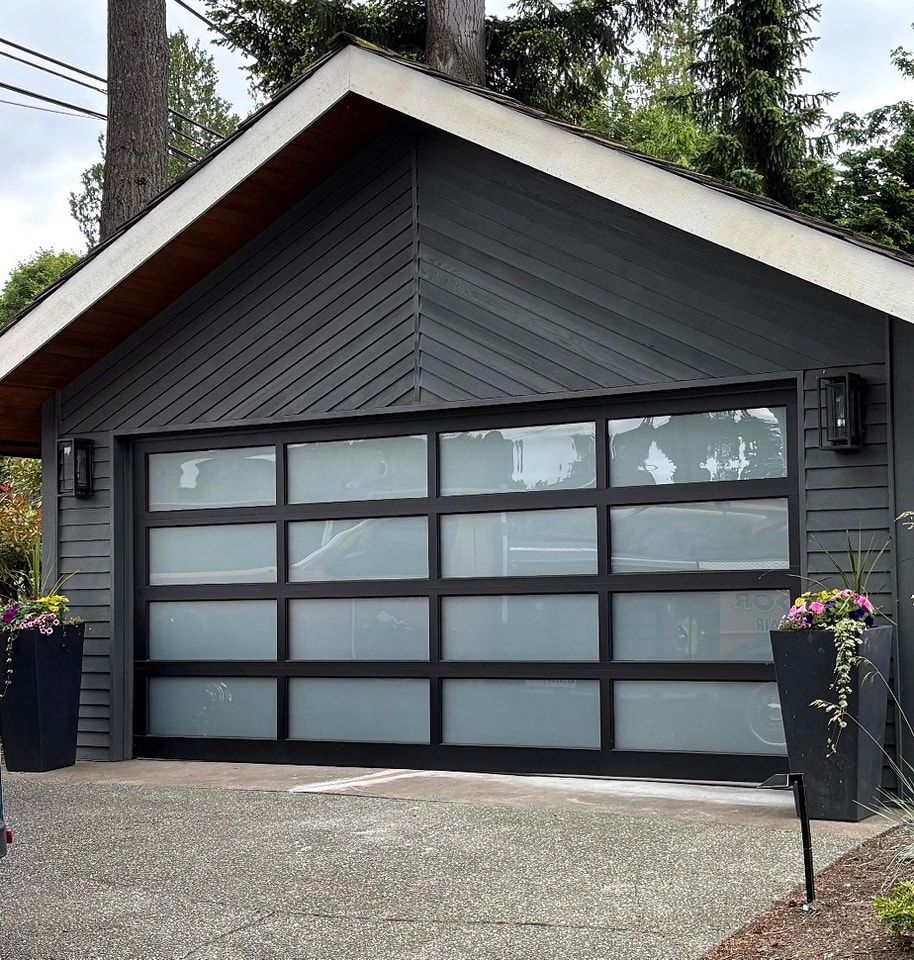 Gray garage with frosted glass door, black trim, and white roof in a wooded driveway