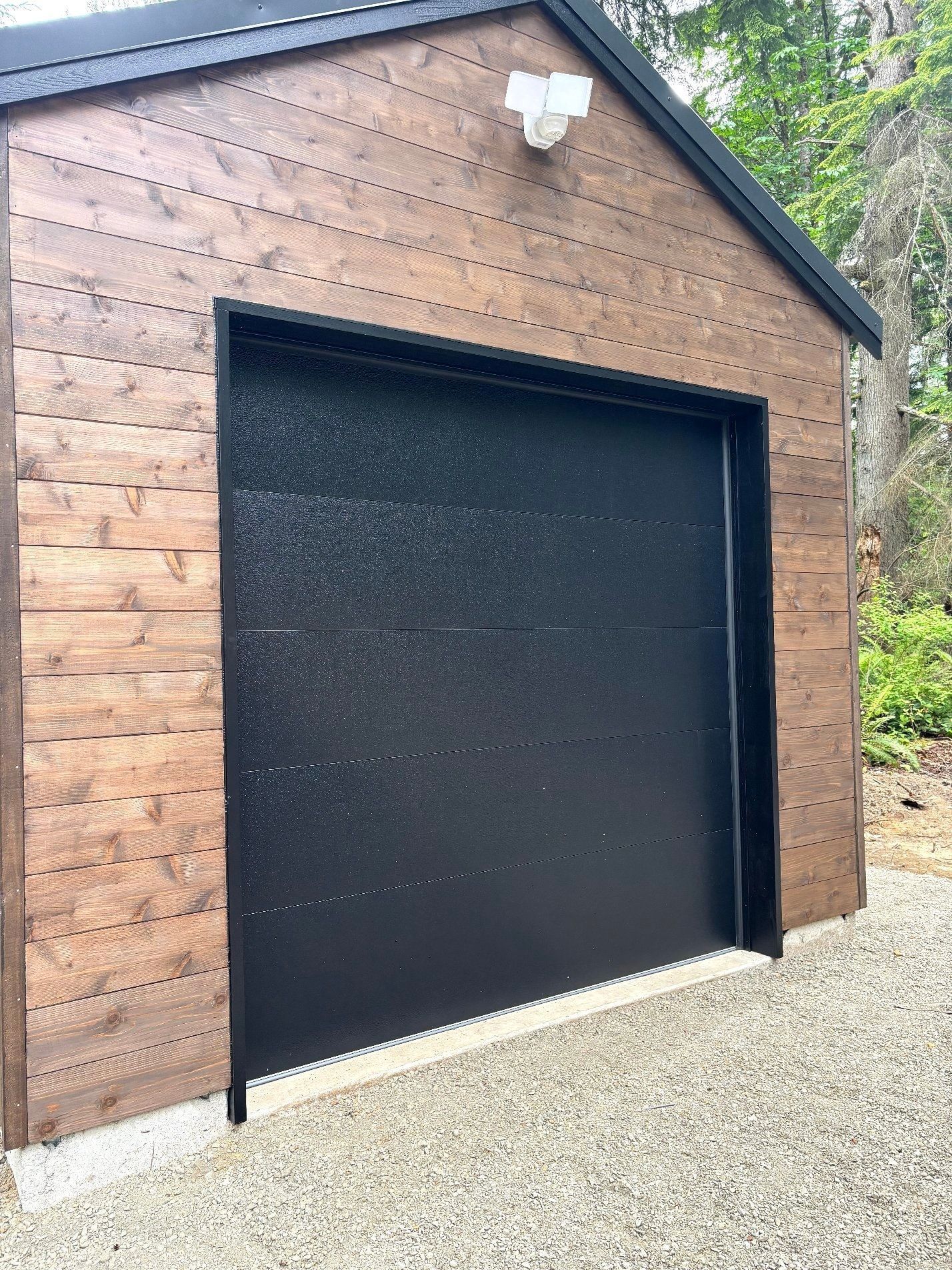 Black garage door on a wooden shed with a gravel driveway and wall light above