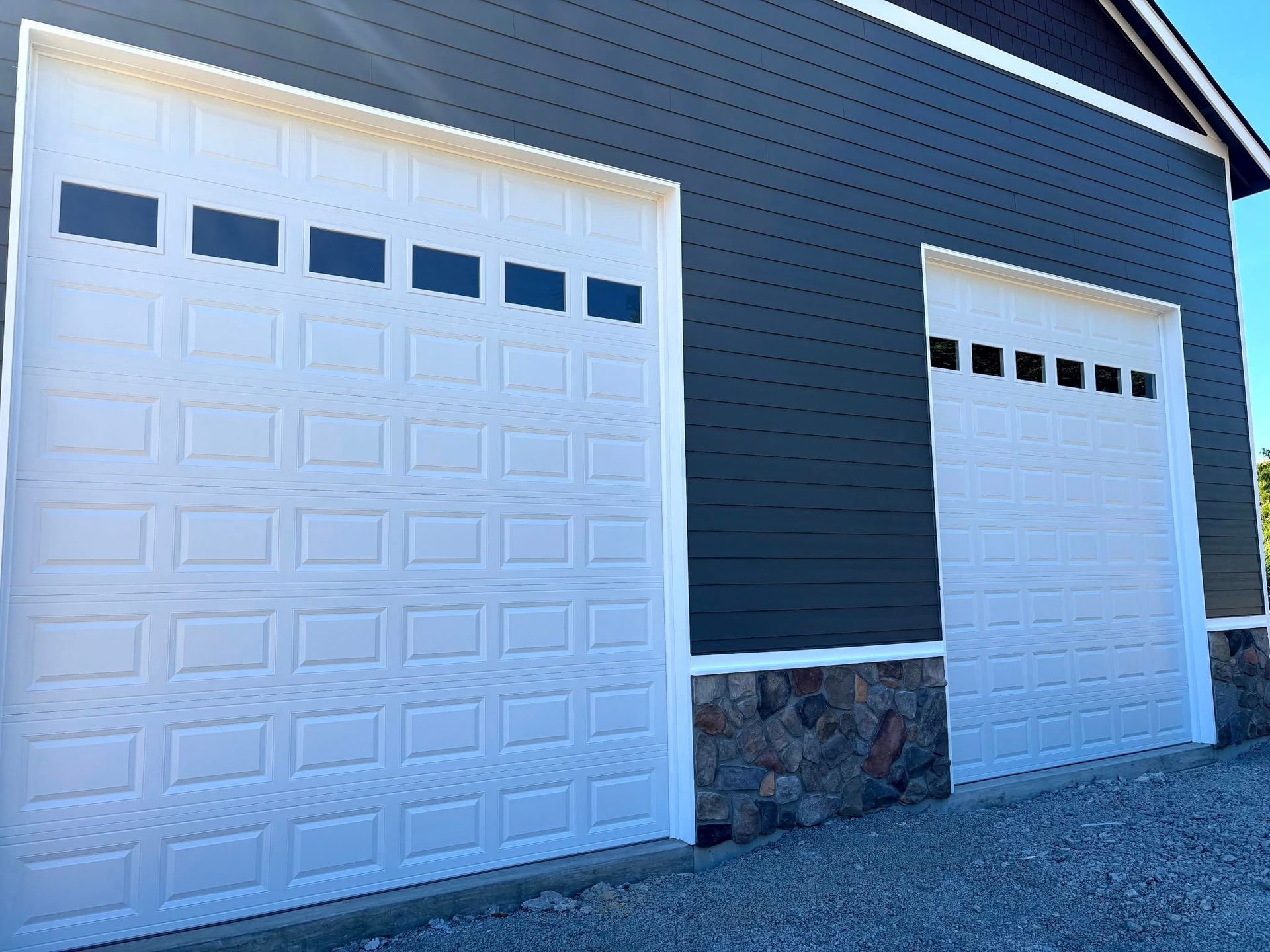Two white garage doors on a dark blue house with a stone base, viewed from an angle.